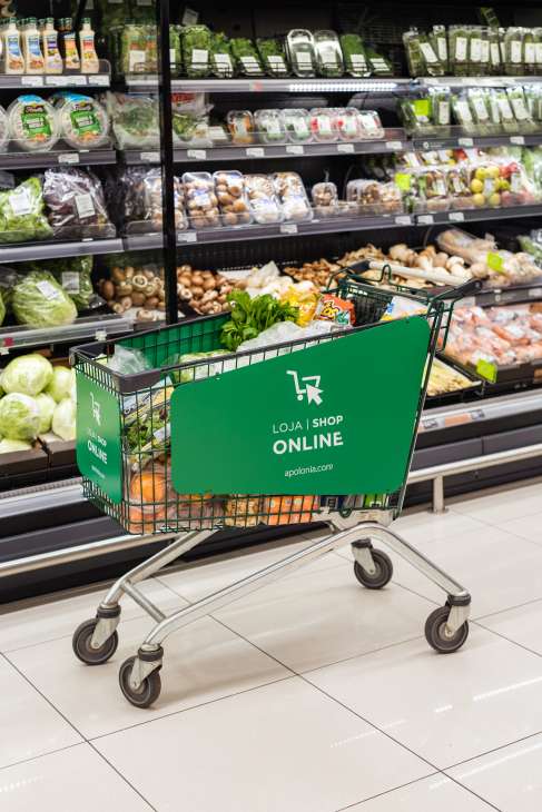 Green shopping cart with Apólonia online grocery store branding in front of refrigerated produce display.