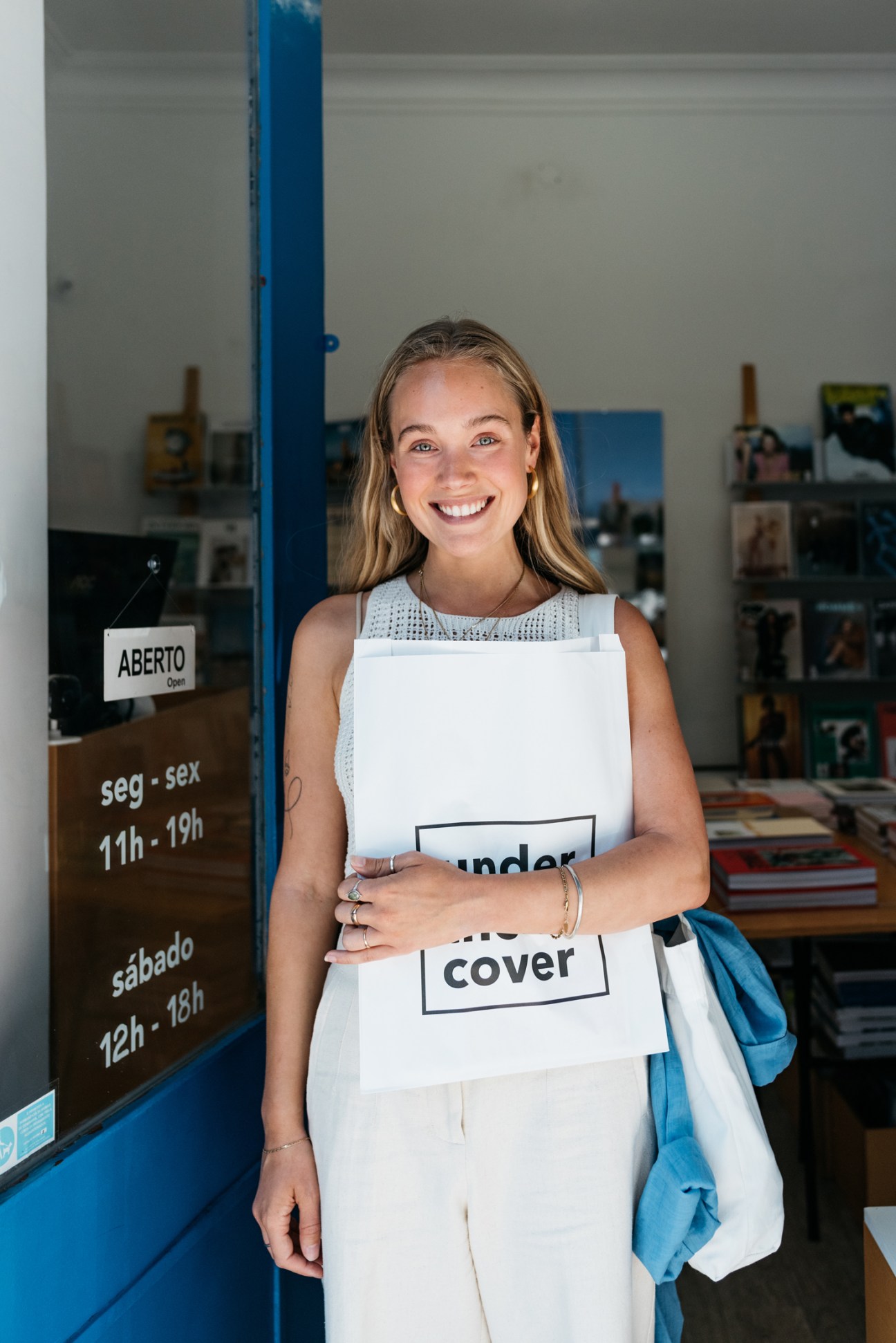 Young blonde woman smiling while holding white shopping bag with 'under cover' text, standing in bookshop