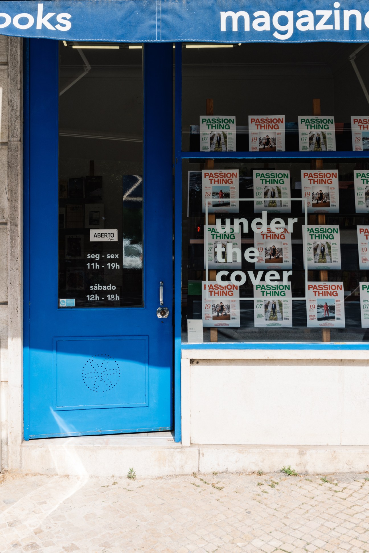 Blue storefront of Under the Cover bookshop in Lisbon with magazines displayed in window and opening hours on door