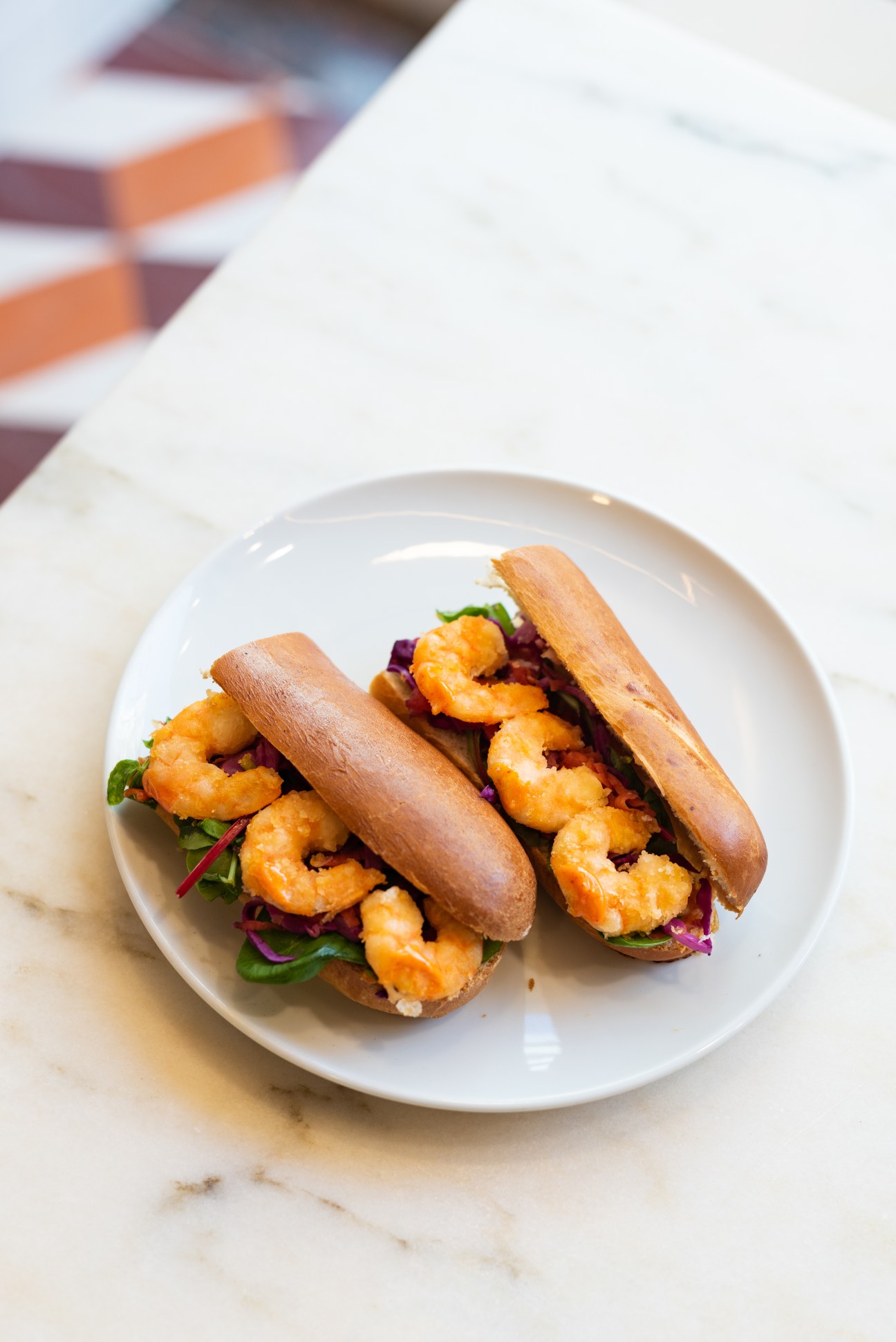 Two shrimp sandwiches with orange shrimp, purple cabbage and greens on brioche buns served on a white plate