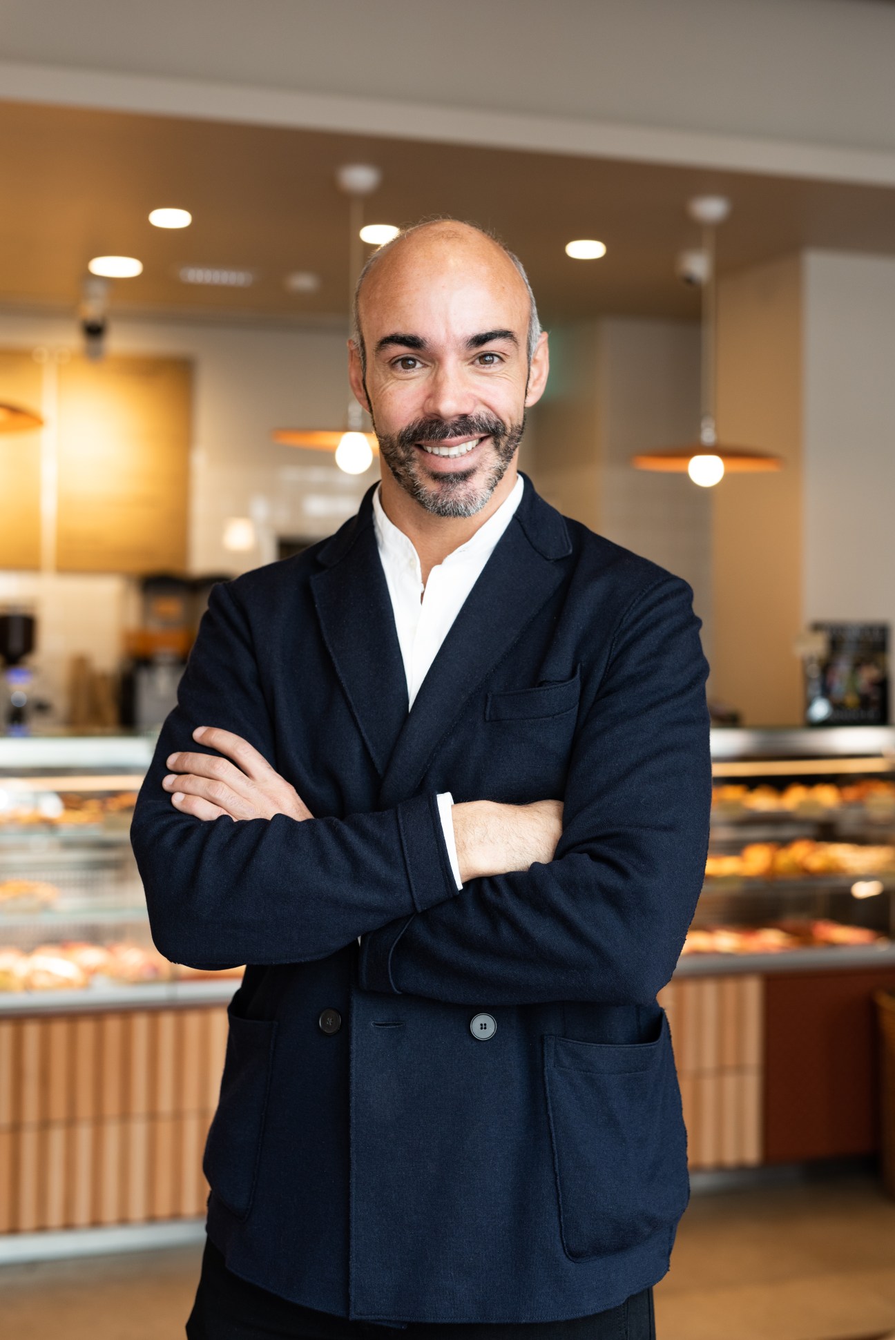 Nuno Carvalho, bald man with beard in dark blazer, arms crossed, smiling in modern bakery interior