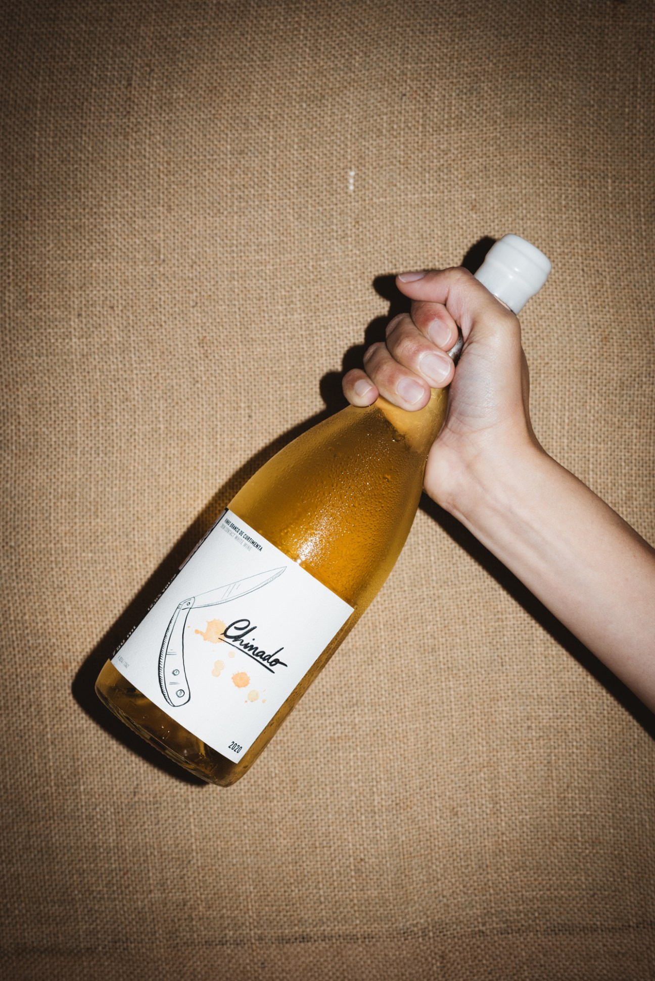 Hand holding amber glass bottle with white label and cork cap against beige textured surface