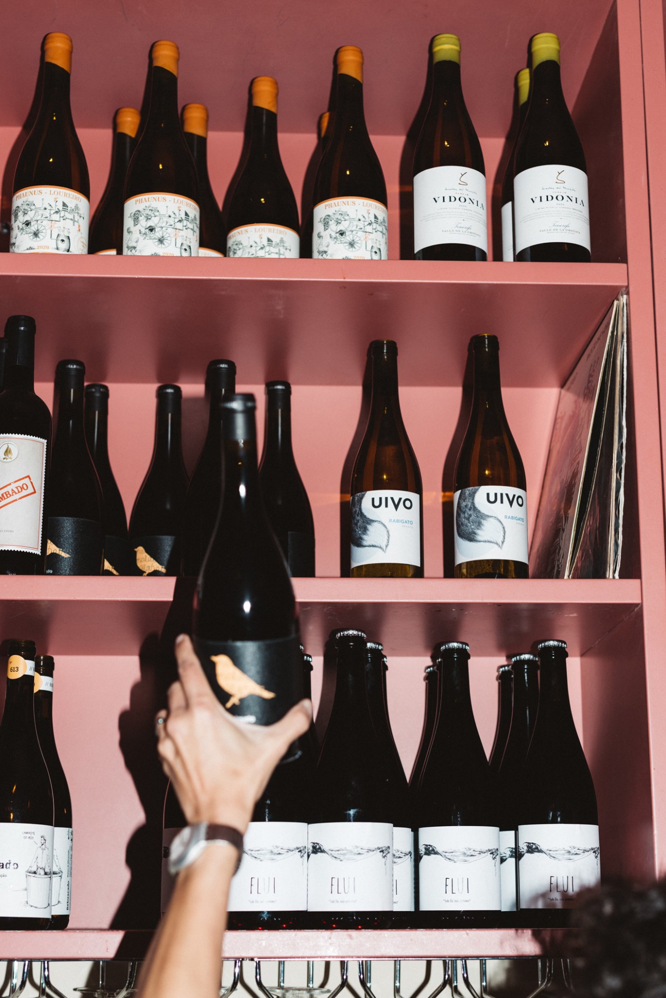 Hand reaching for wine bottle on pink shelves displaying various wine bottles including Vidonia and Uivo labels