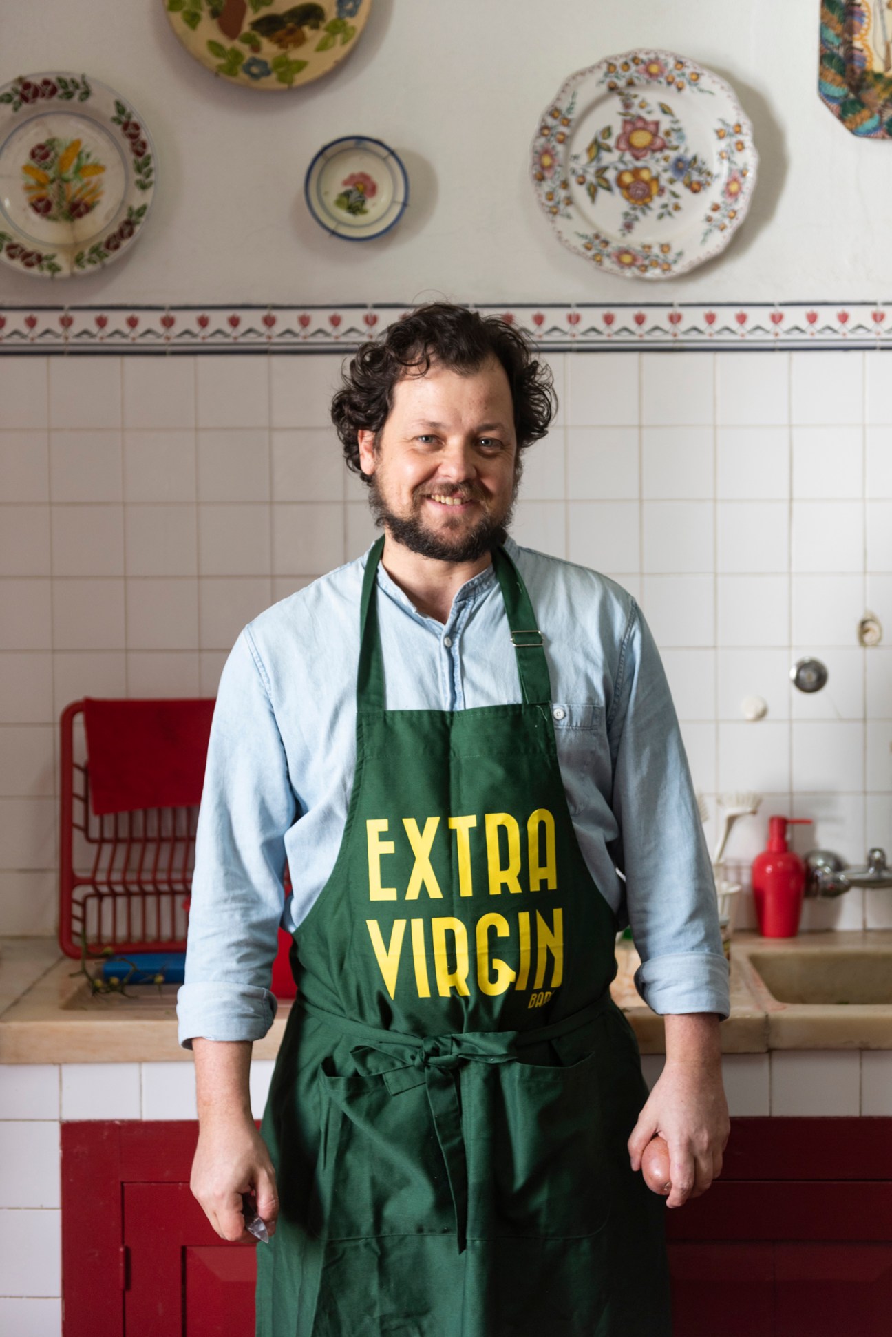 Chef João Rodrigues wearing green apron with 'EXTRA VIRGIN' text in kitchen with decorative plates on wall