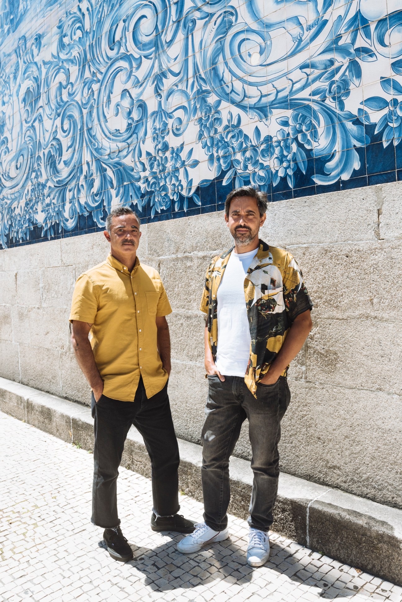 Two men standing against a wall with blue and white ceramic tile artwork, one wearing yellow shirt and dark pants, the other wearing patterned shirt and jeans.