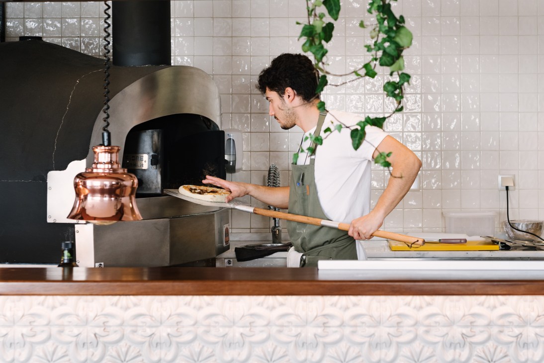 Chef using long wooden peel to remove pizza from wood-fired oven in tiled kitchen at Casa do Gadanha