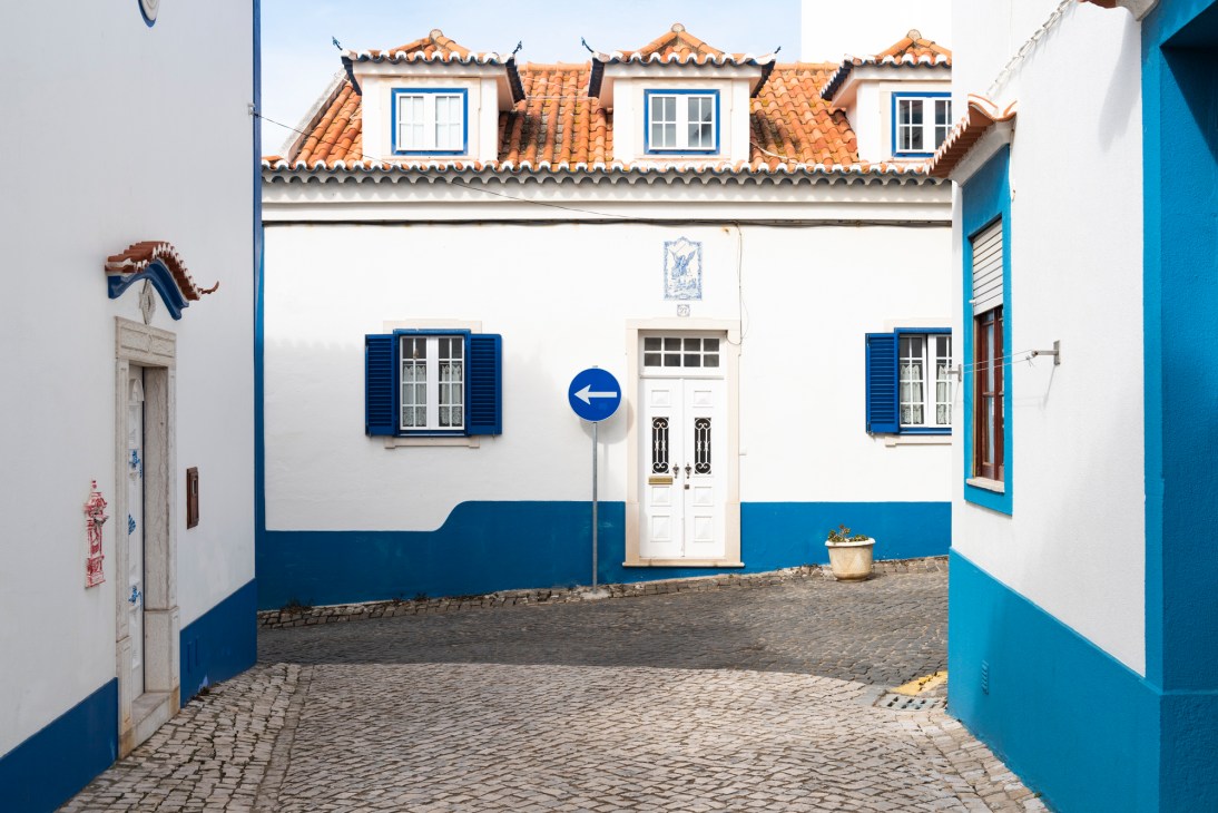 Traditional Portuguese buildings with white walls, blue shutters and doors, and terracotta tile roofs lining a cobblestone street in Ericeira.