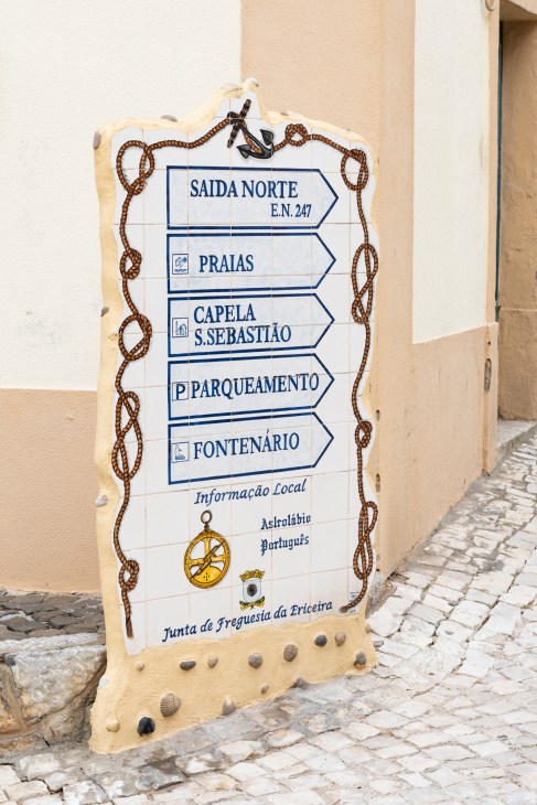 A directional sign in Ericeira with nautical rope decoration pointing to beaches, chapels, and local amenities in Portuguese.