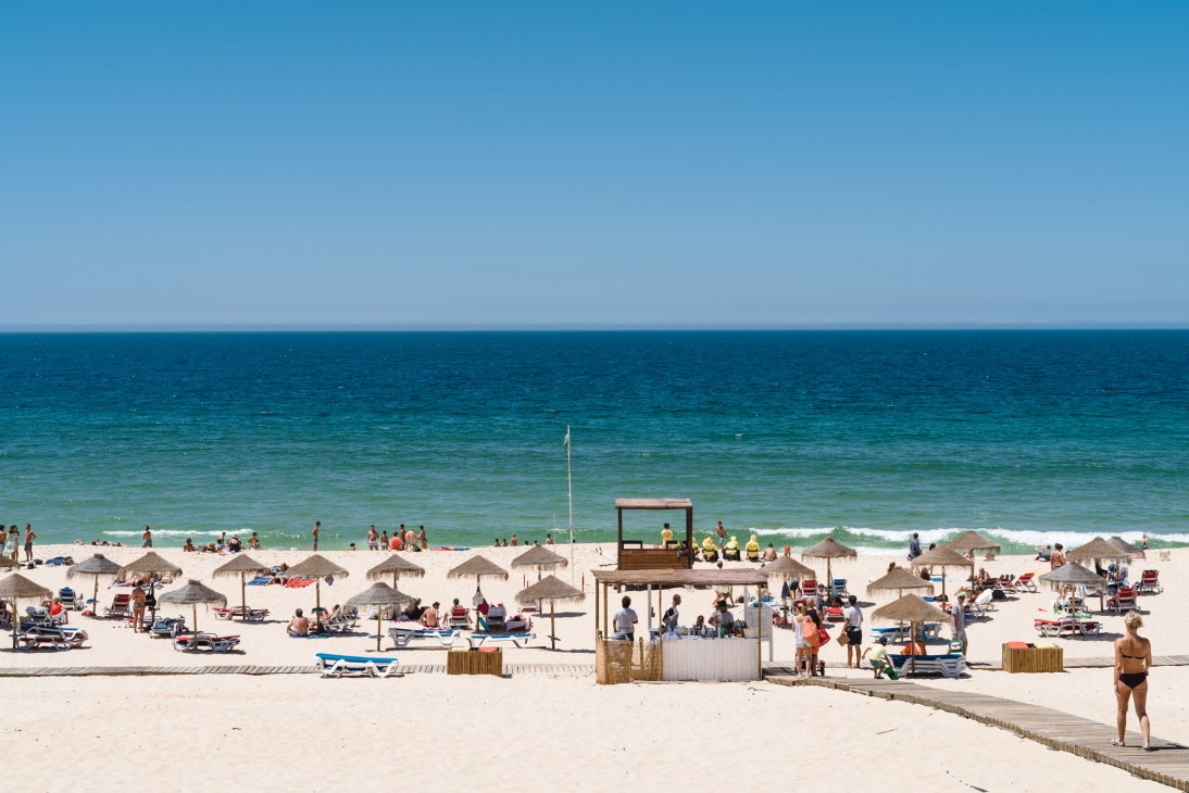 Busy beach scene with straw umbrellas, sunbathers, and a wooden beach bar structure at Pego beach in Comporta, Portugal.