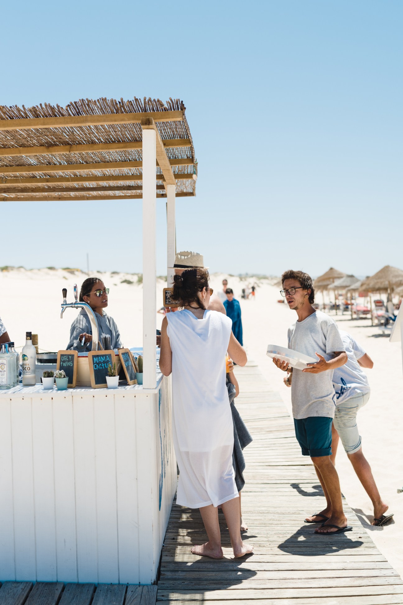 Customers ordering at a beachside food counter with white walls and thatched roof shade structure.