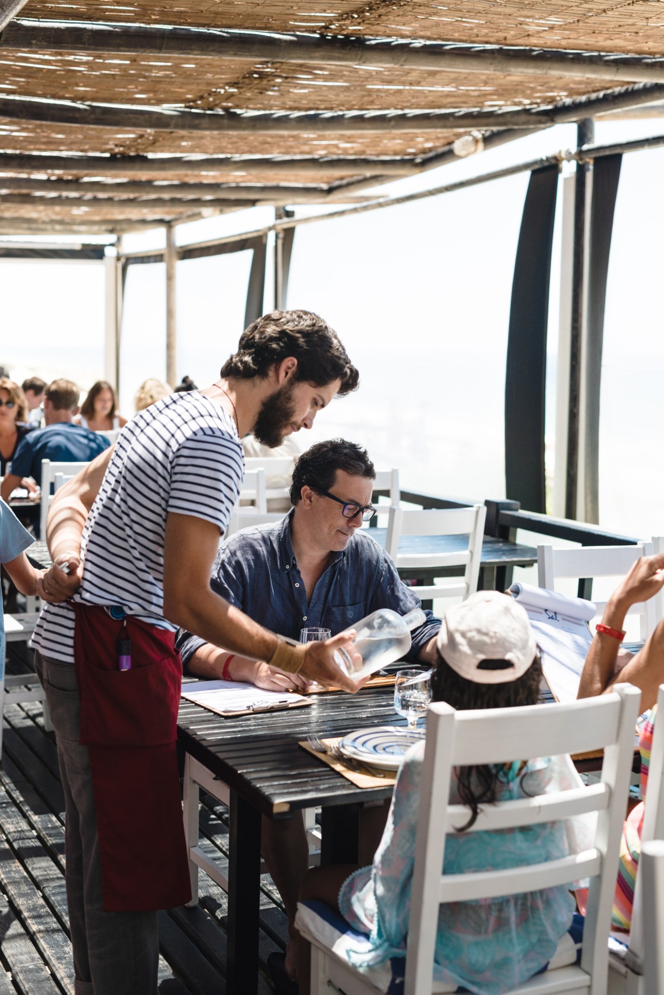 A waiter in striped shirt serves customers dining at a beachside restaurant terrace with wooden pergola.