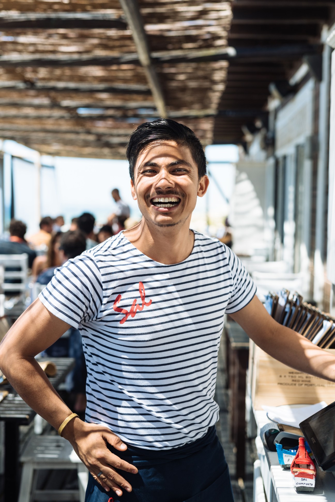 Bikash Baral, a waiter at Sal beach restaurant, smiling at the camera wearing a striped shirt with the Sal logo.