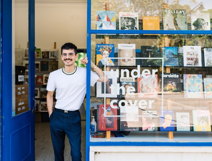 Smiling man with mustache in white t-shirt and dark pants standing in doorway of bookshop with blue frame