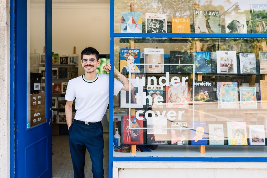 Smiling man with mustache in white t-shirt and dark pants standing in doorway of bookshop with blue frame