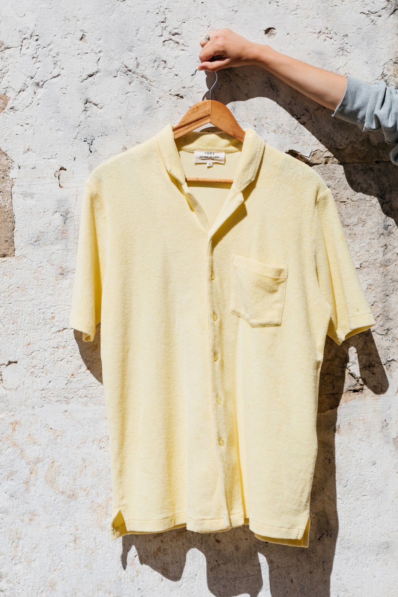 Person holding yellow button-up shirt on wooden hanger against weathered white wall