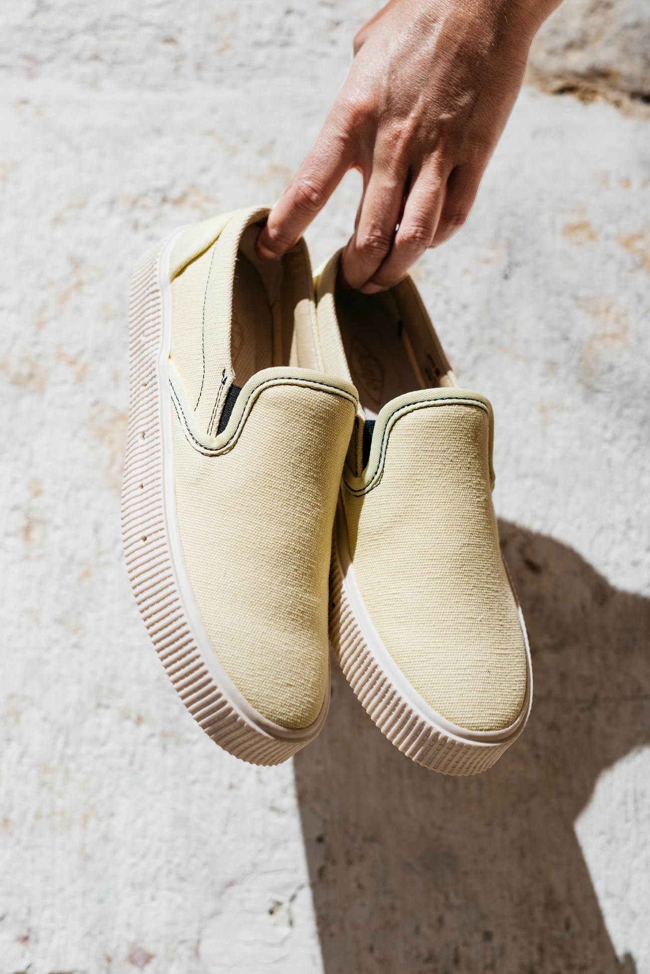 Hand holding a pair of beige canvas slip-on sneakers with ridged white soles against a light stone surface