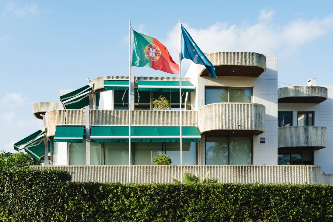 Modern white apartment building with curved balconies, green awnings, and Portuguese and EU flags flying on flagpoles