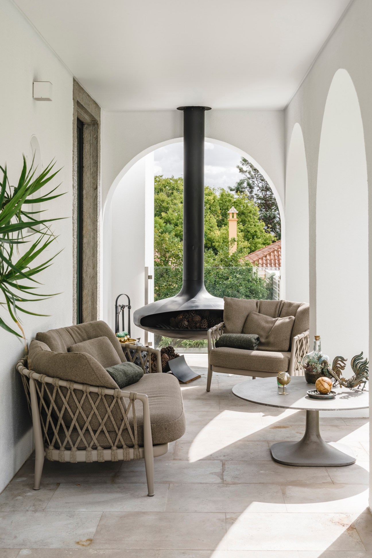 Modern covered terrace with suspended fireplace, neutral armchairs, and arched opening overlooking greenery