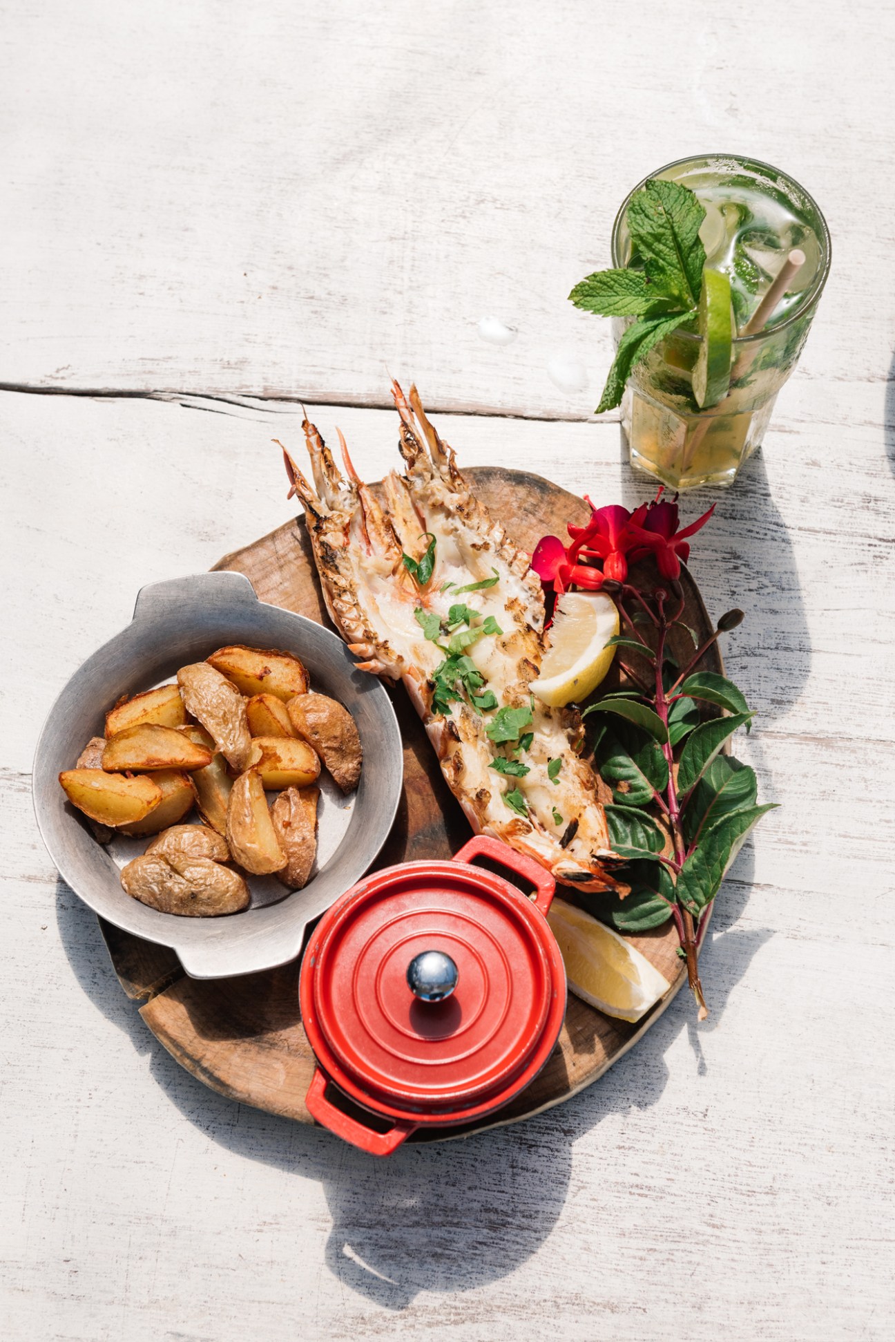 Grilled tiger prawn with herbs and lemon on a wooden board, served with fried potatoes, a mojito cocktail, and red flower garnish on white wood.