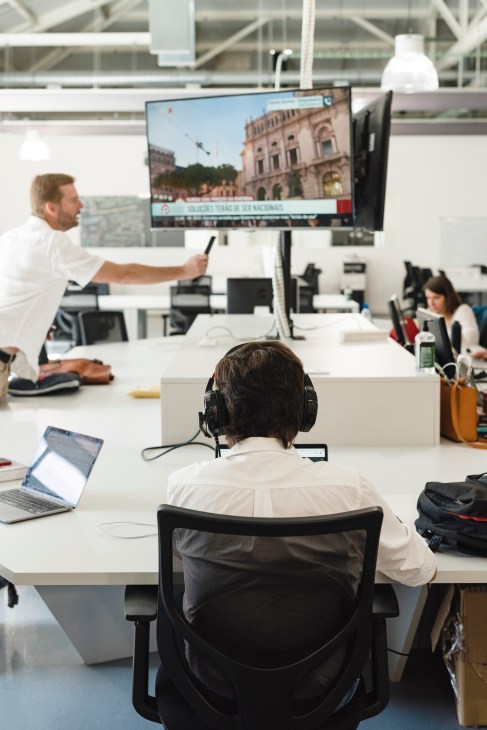 Staff members work at desks in a modern newsroom while a man points at a large monitor displaying a Portuguese building.
