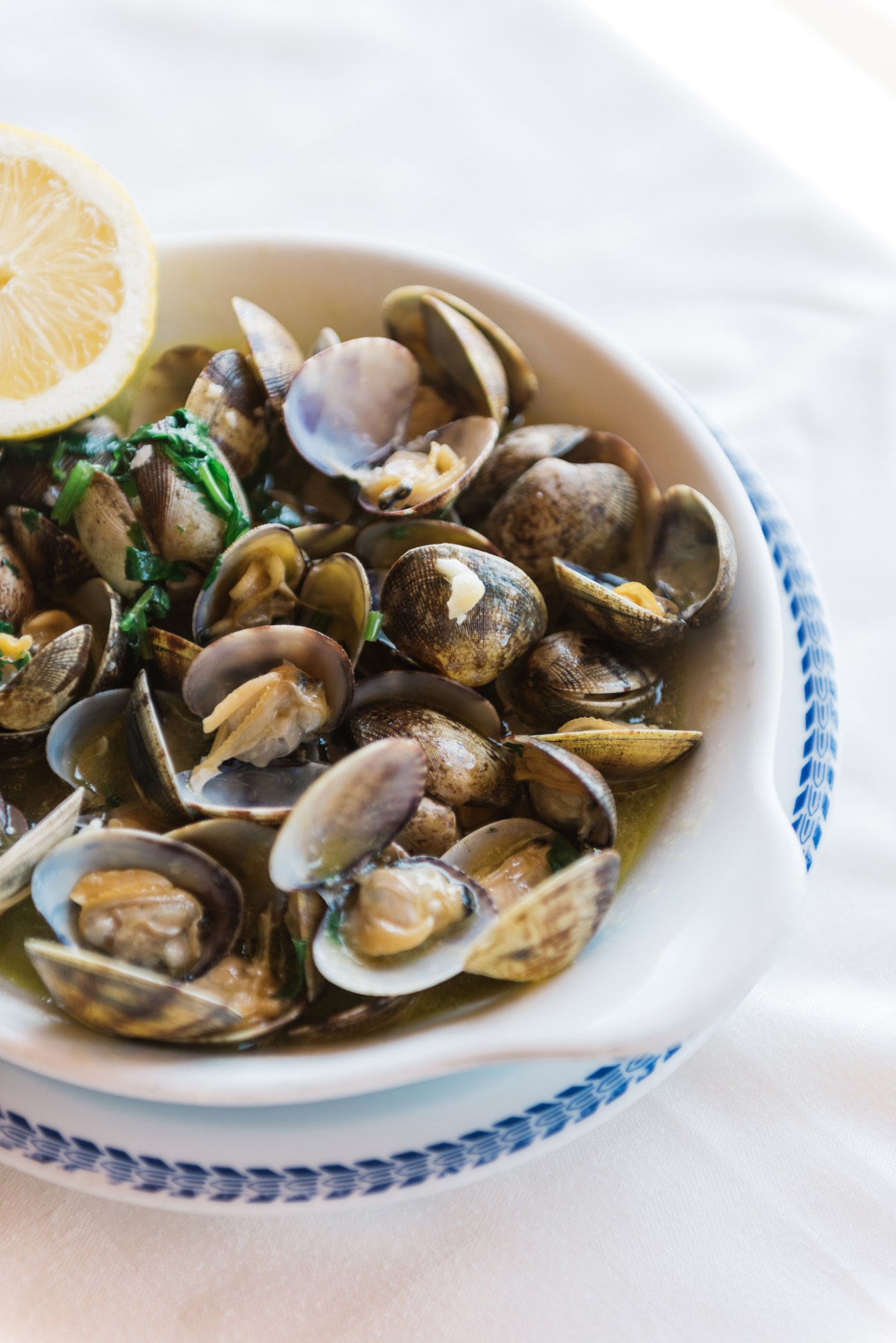 A white bowl with blue trim filled with clams garnished with parsley and lemon wedge.