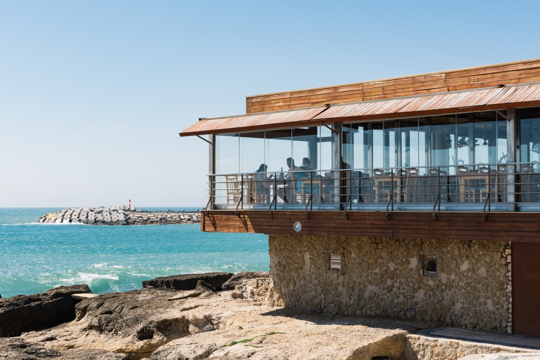 Modern wooden beach restaurant with glass windows overlooking turquoise ocean and rocky coastline.