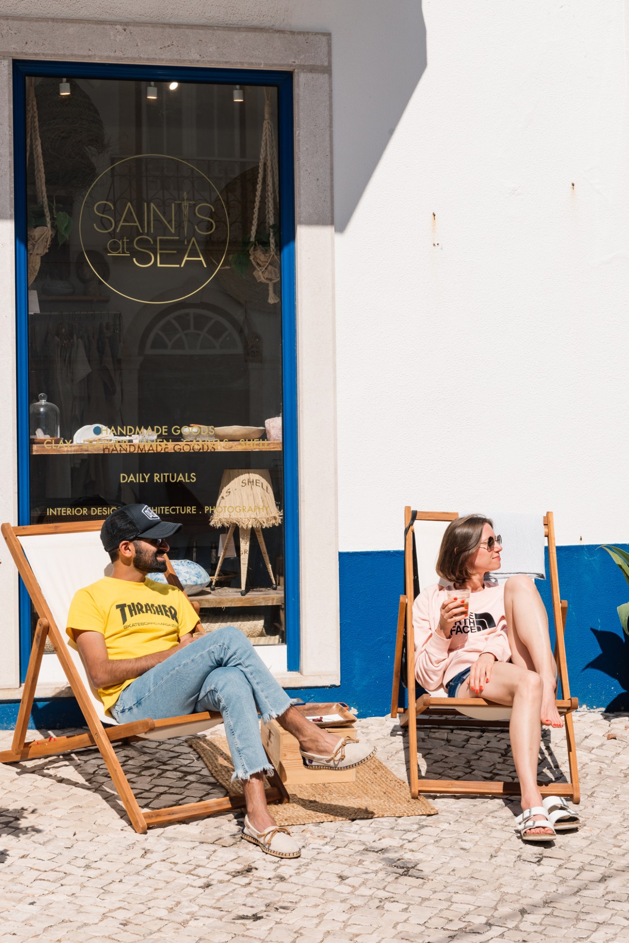 Two people sitting in wooden deck chairs outside a blue storefront with 'Saints at Sea' signage.