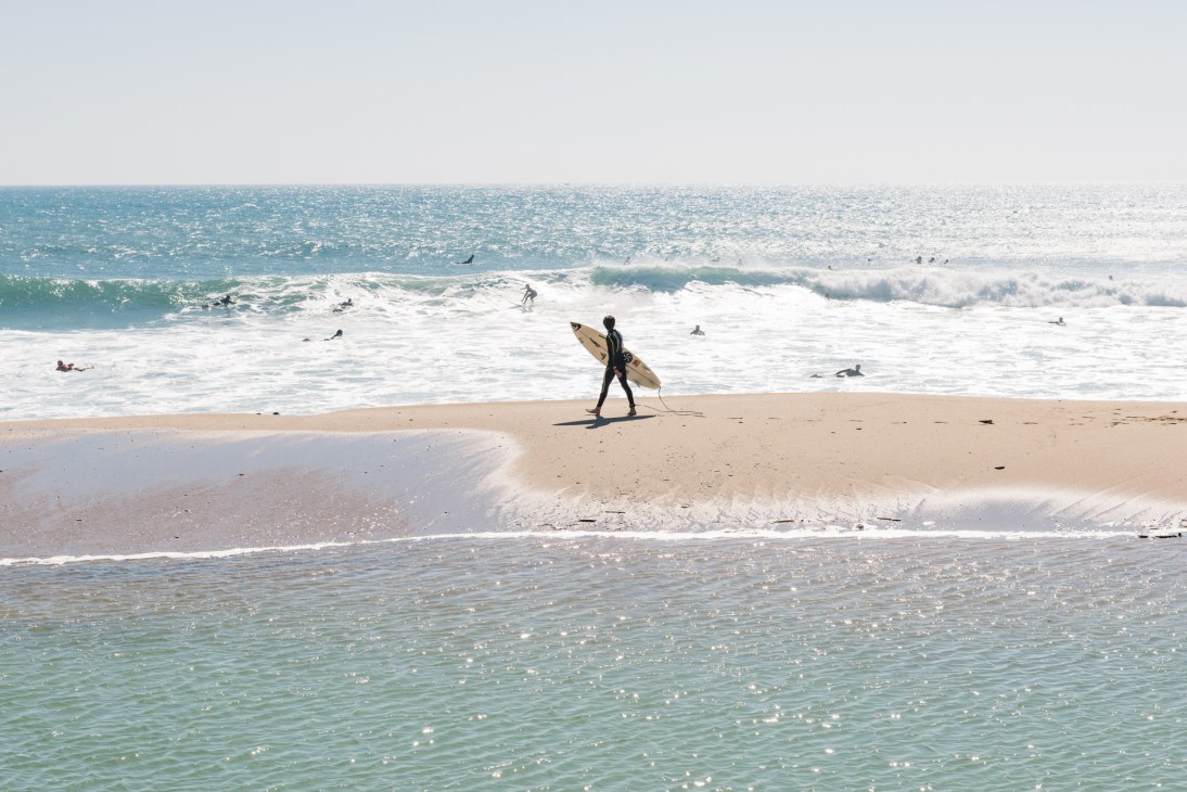 Surfer carrying a wooden board walking on a sandy beach with waves and other surfers in the water.