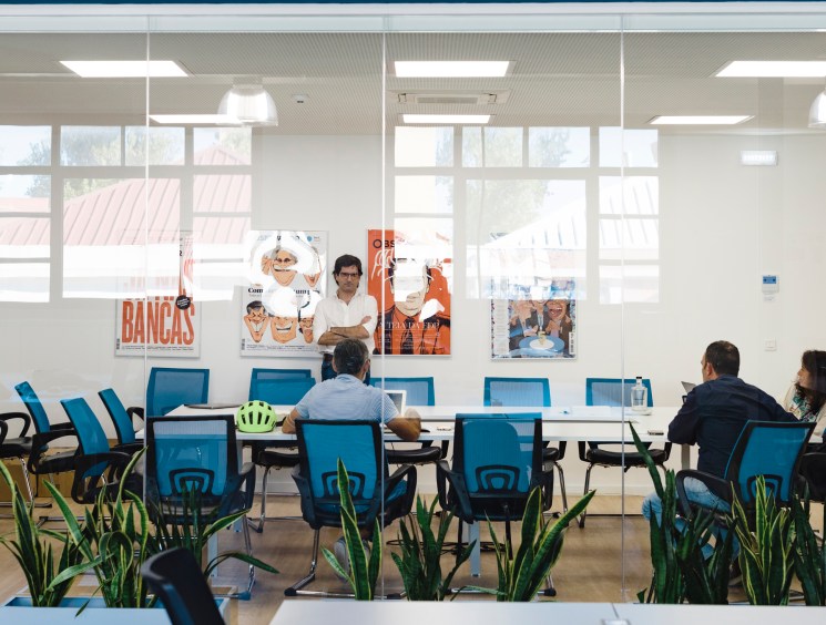Observador staff members meet at a conference table in a bright, modern office with blue chairs and magazine covers on the wall.