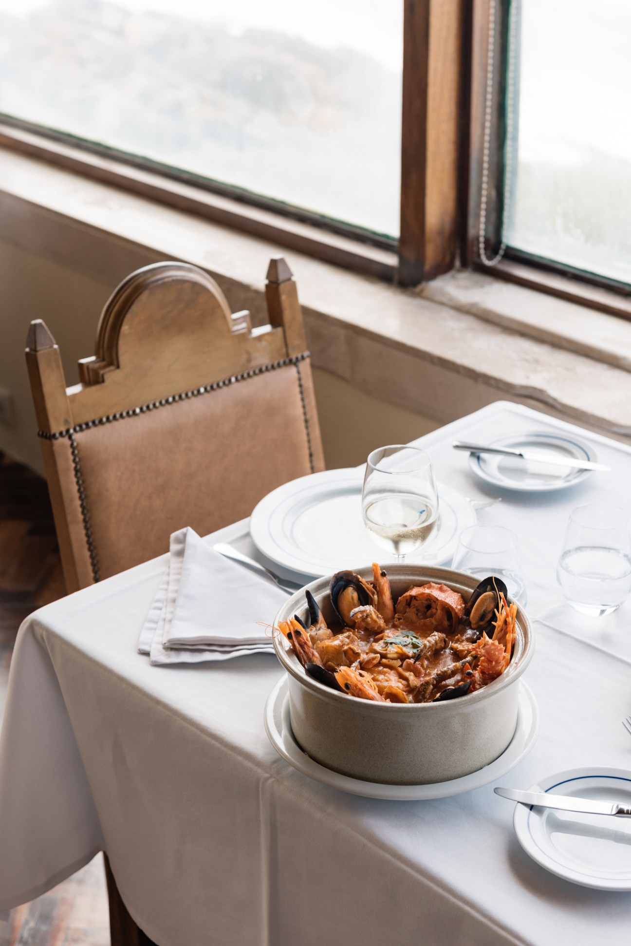 Table set with white linens, plate settings, and a bowl of grilled shellfish beside a window overlooking the ocean.