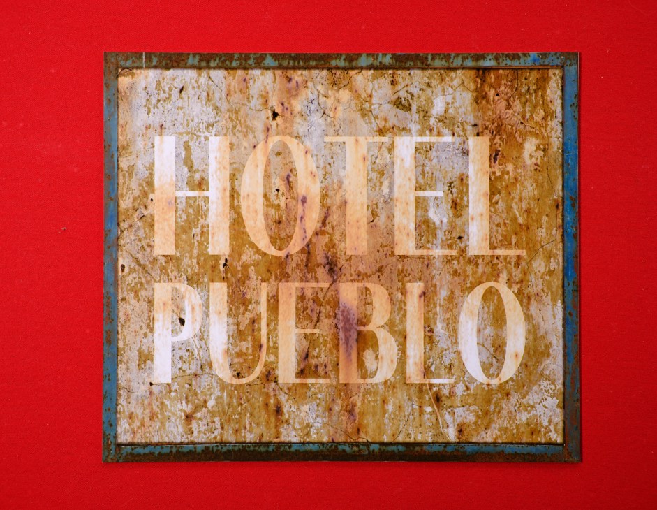 Weathered hotel sign reading 'HOTEL PUEBLO' with aged, distressed lettering on worn surface