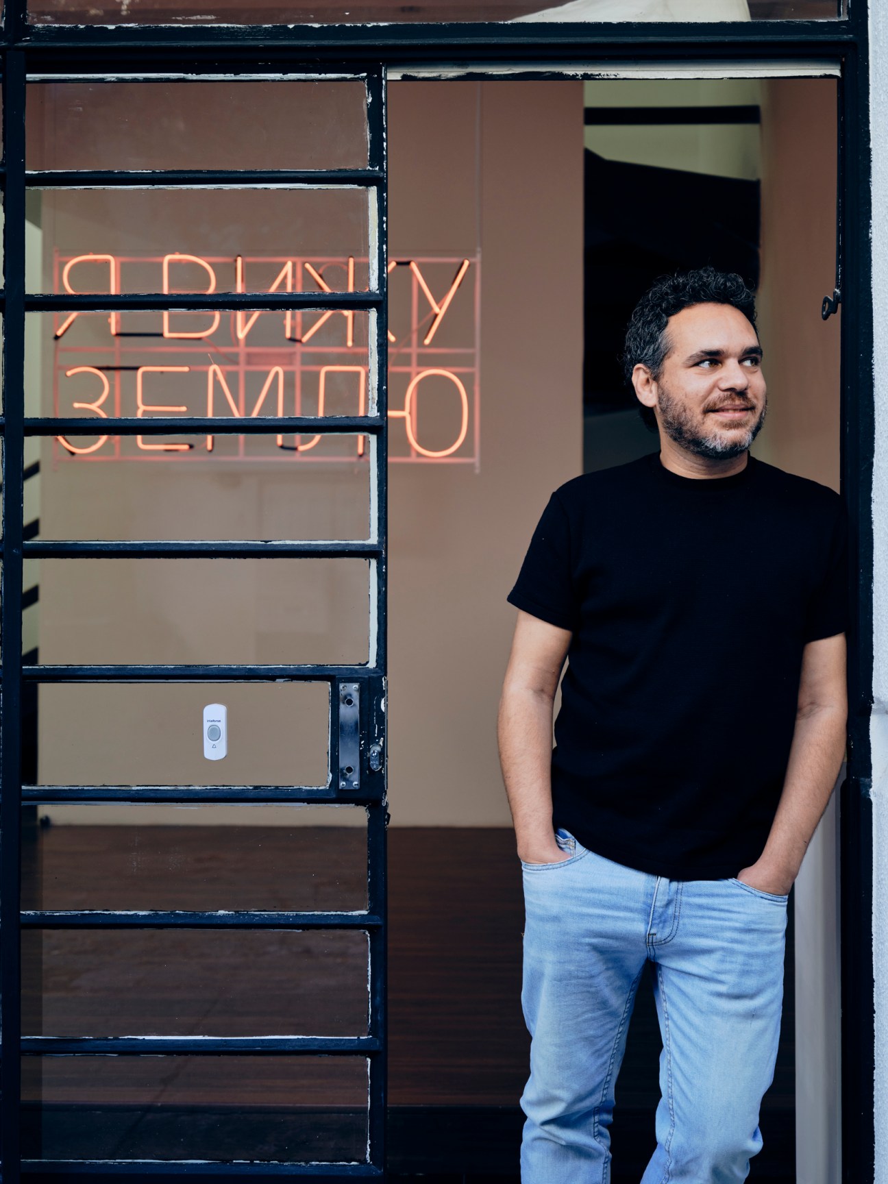 Man in black t-shirt and jeans standing in doorway with orange neon sign visible through window