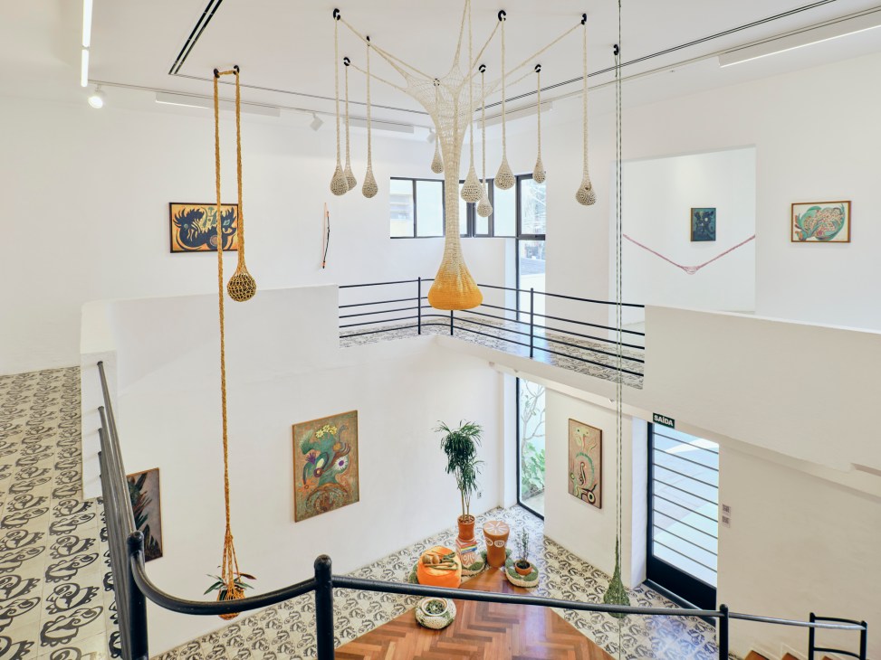 Modern gallery interior with double-height ceiling, hanging woven sculptures, artwork on walls, and geometric railings