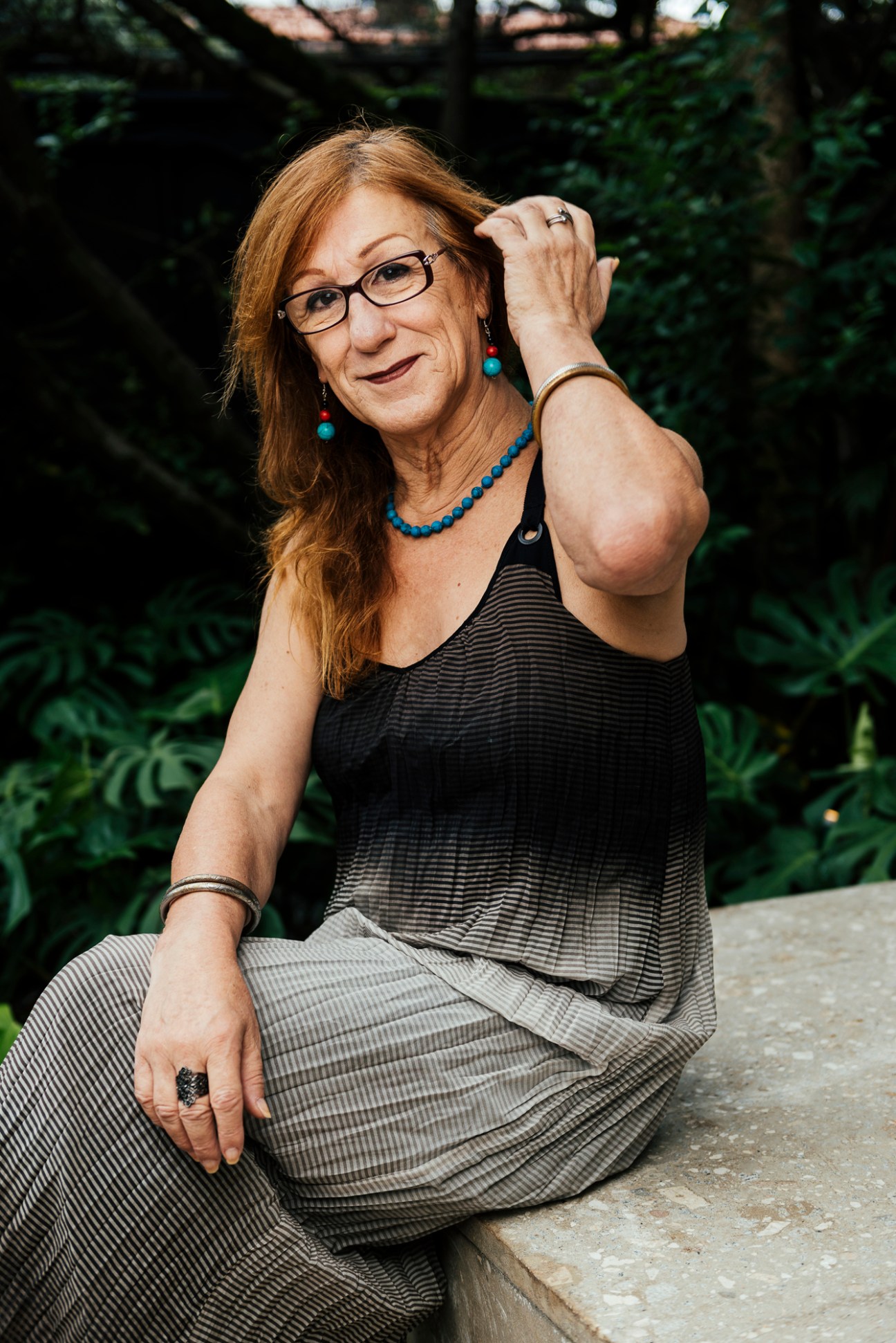 Laerte, a Brazilian cartoonist, sitting outdoors wearing glasses and a blue beaded necklace