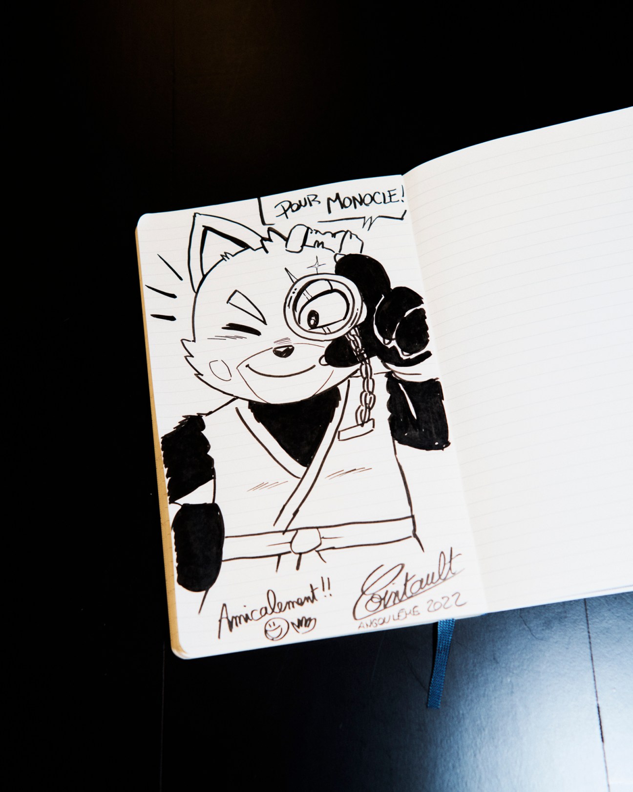 Hand-drawn manga character in a notebook with monocle saying 'Pour Monocle!' signed by artist