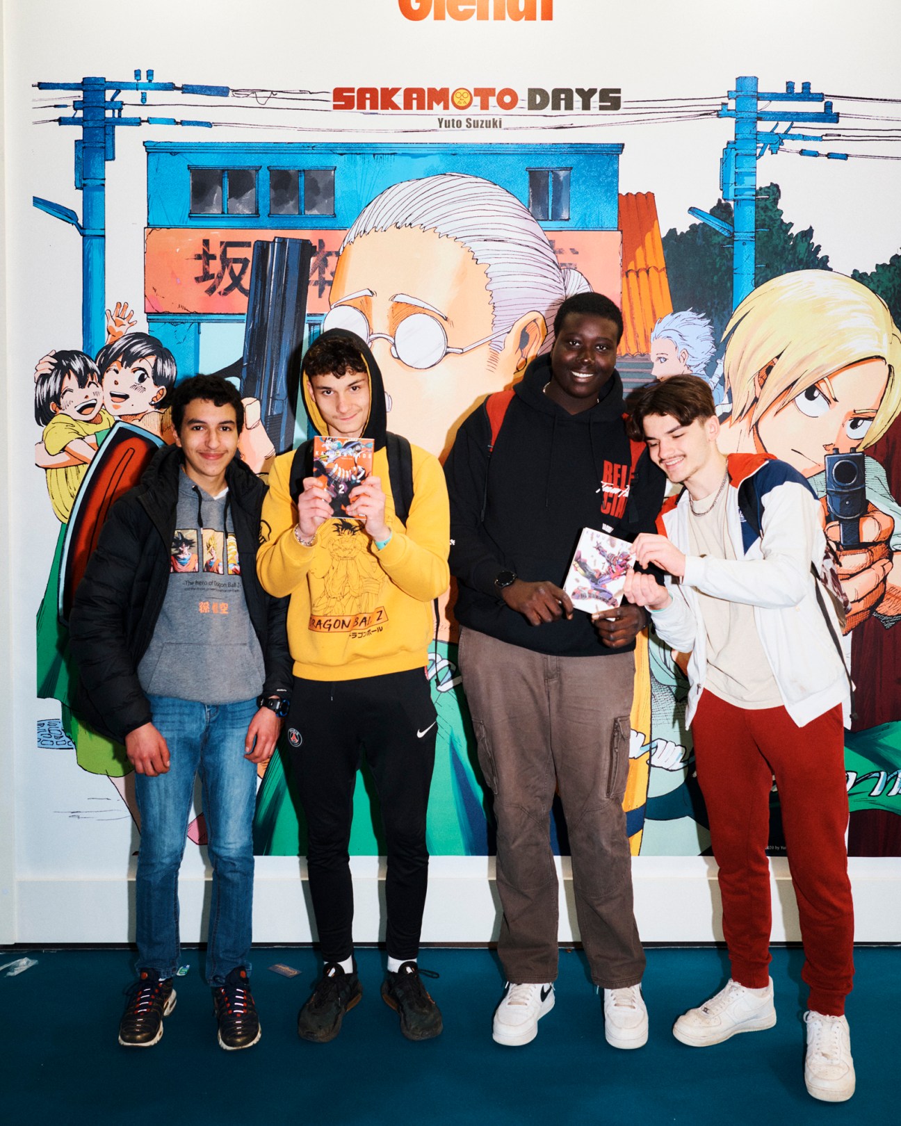 Four young people posing with manga books in front of a Sakamoto Days anime poster at Angoulême festival