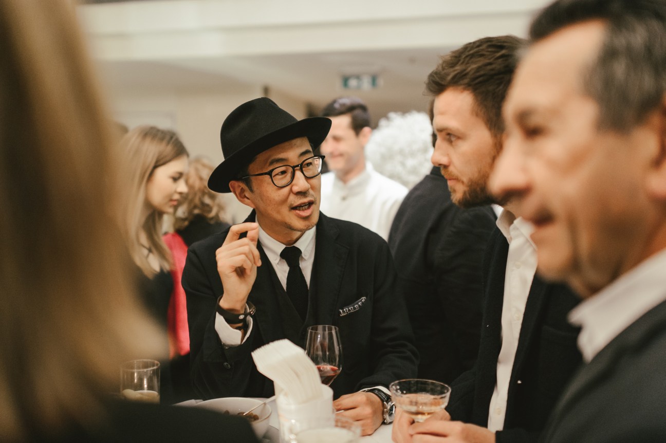 Well-dressed man in black hat and glasses speaking at a conference table with wine glasses