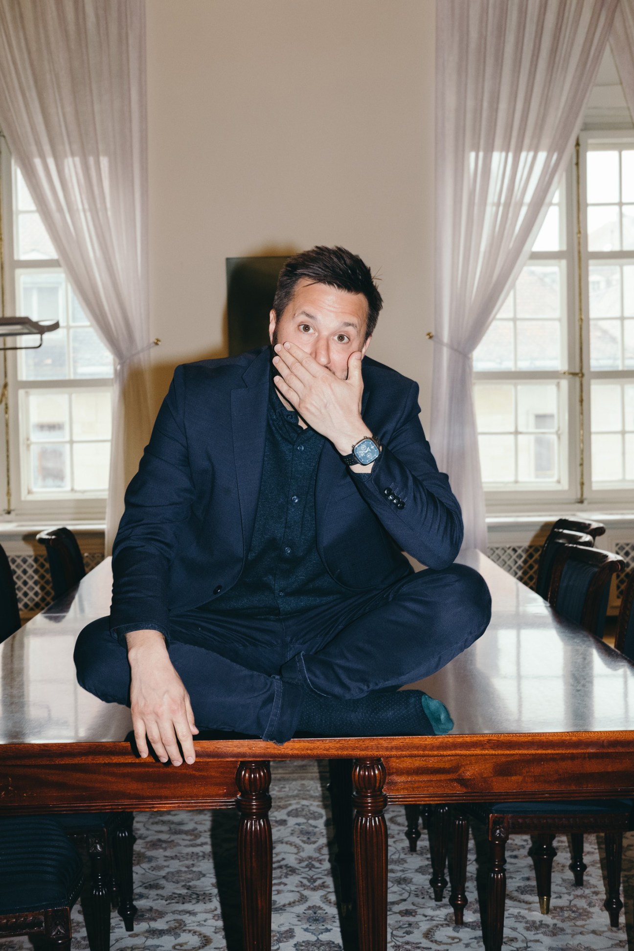 Man in dark navy suit sitting on wooden table in elegant room with tall windows and sheer curtains, hand near mouth in thoughtful pose.