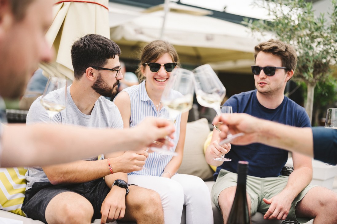 Four people toasting with white wine glasses outdoors at a sunny terrace gathering