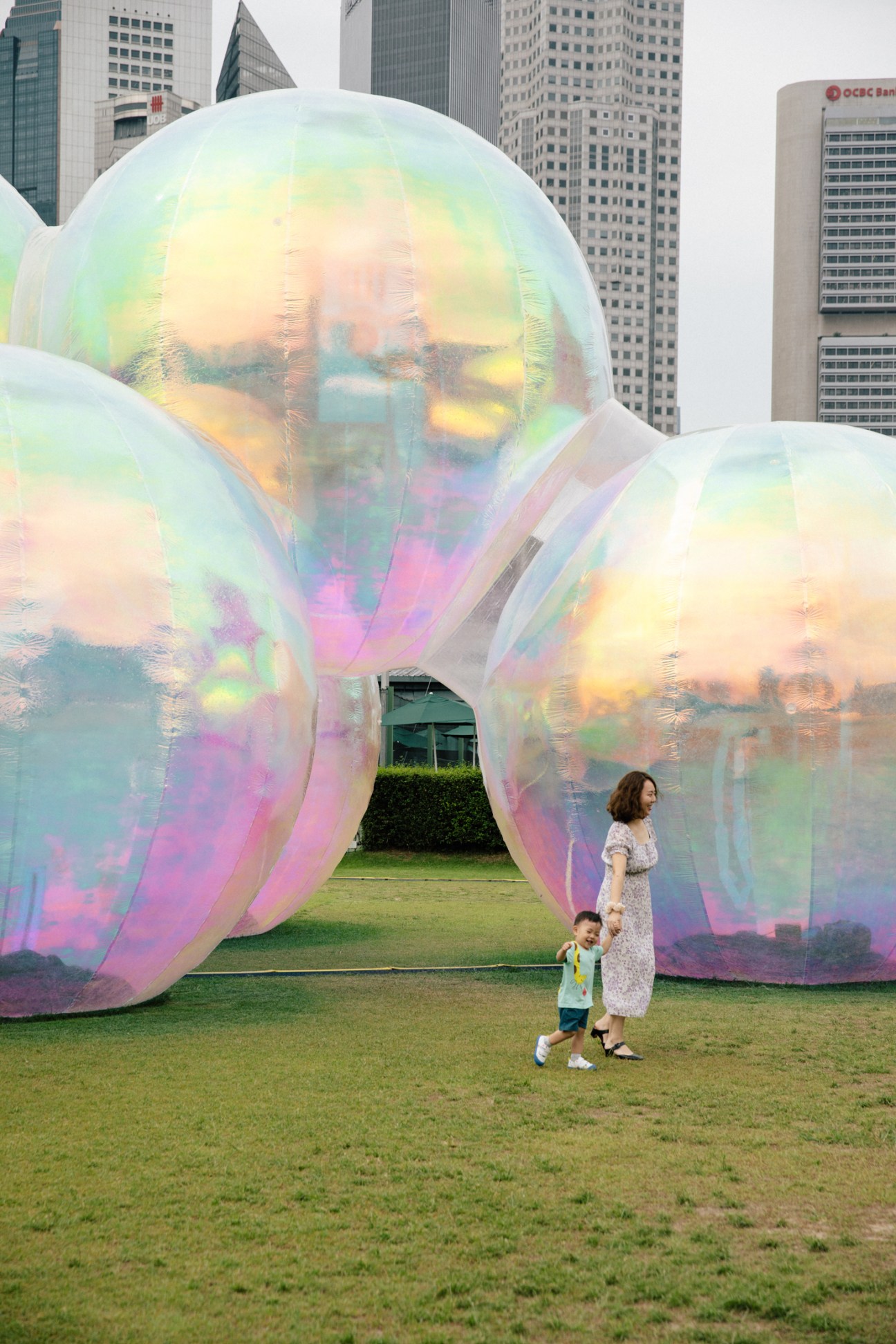Large iridescent bubble-like art installation with rainbow reflections on grass with woman and child walking nearby