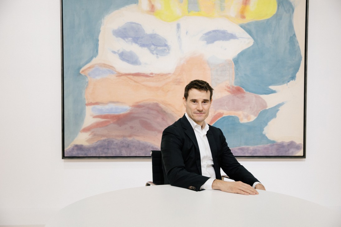 Nick Simunovic sits at a white table in front of a large abstract painting with colorful clouds