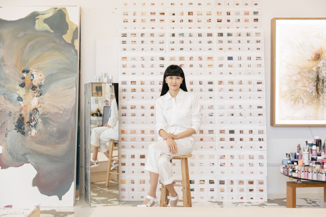 Dawn Ng sits on a wooden stool in her art studio surrounded by abstract paintings and color samples on the walls