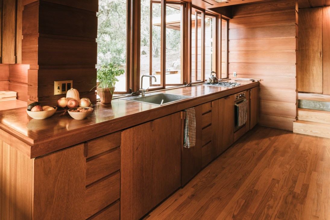 Modern wooden kitchen with island counter, stainless steel sink, and large windows overlooking foothills.