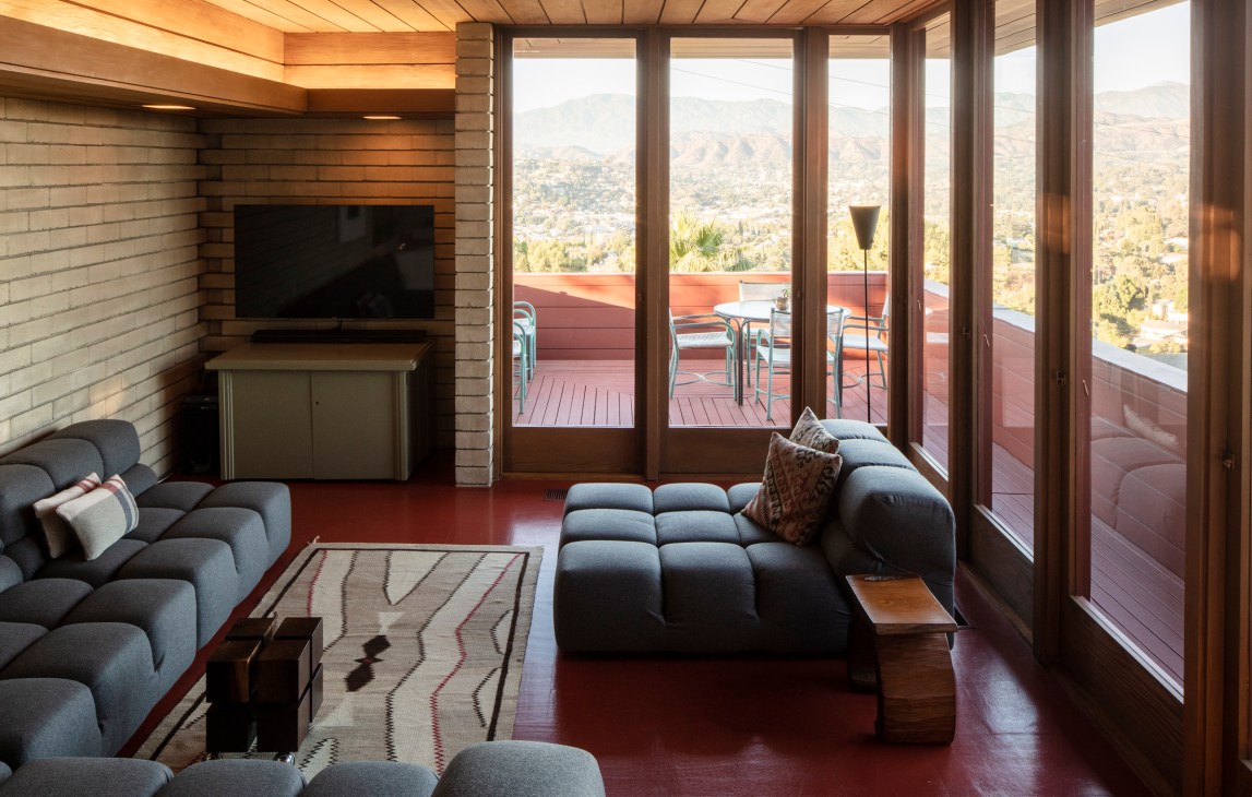 Modern living room with panoramic glass doors overlooking foothills, featuring dark upholstered seating and minimalist furnishings.
