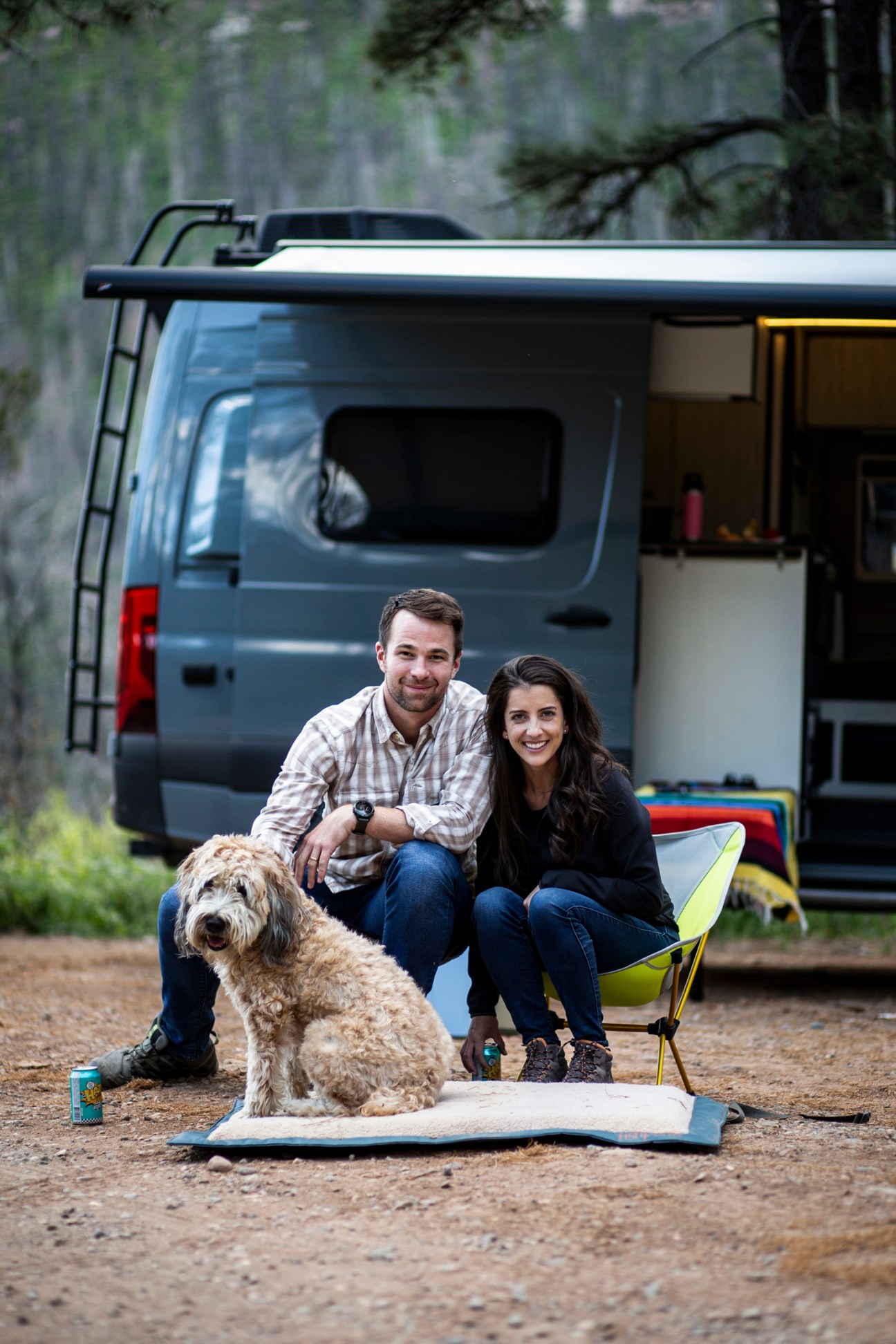A couple with their dog sitting outside a gray camper van in a wooded area
