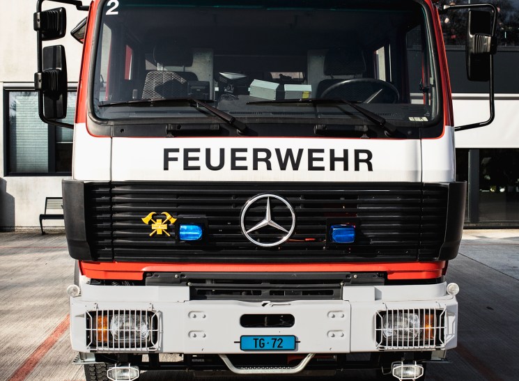 Red and white Mercedes-Benz fire truck with FEUERWEHR marking and blue emergency lights parked outside building