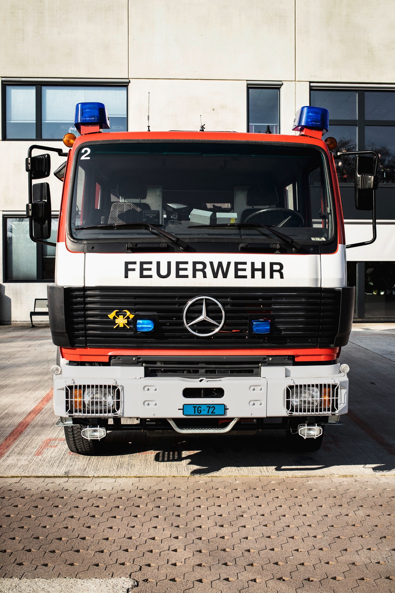 Red and white Mercedes-Benz fire truck with FEUERWEHR marking and blue emergency lights parked outside building