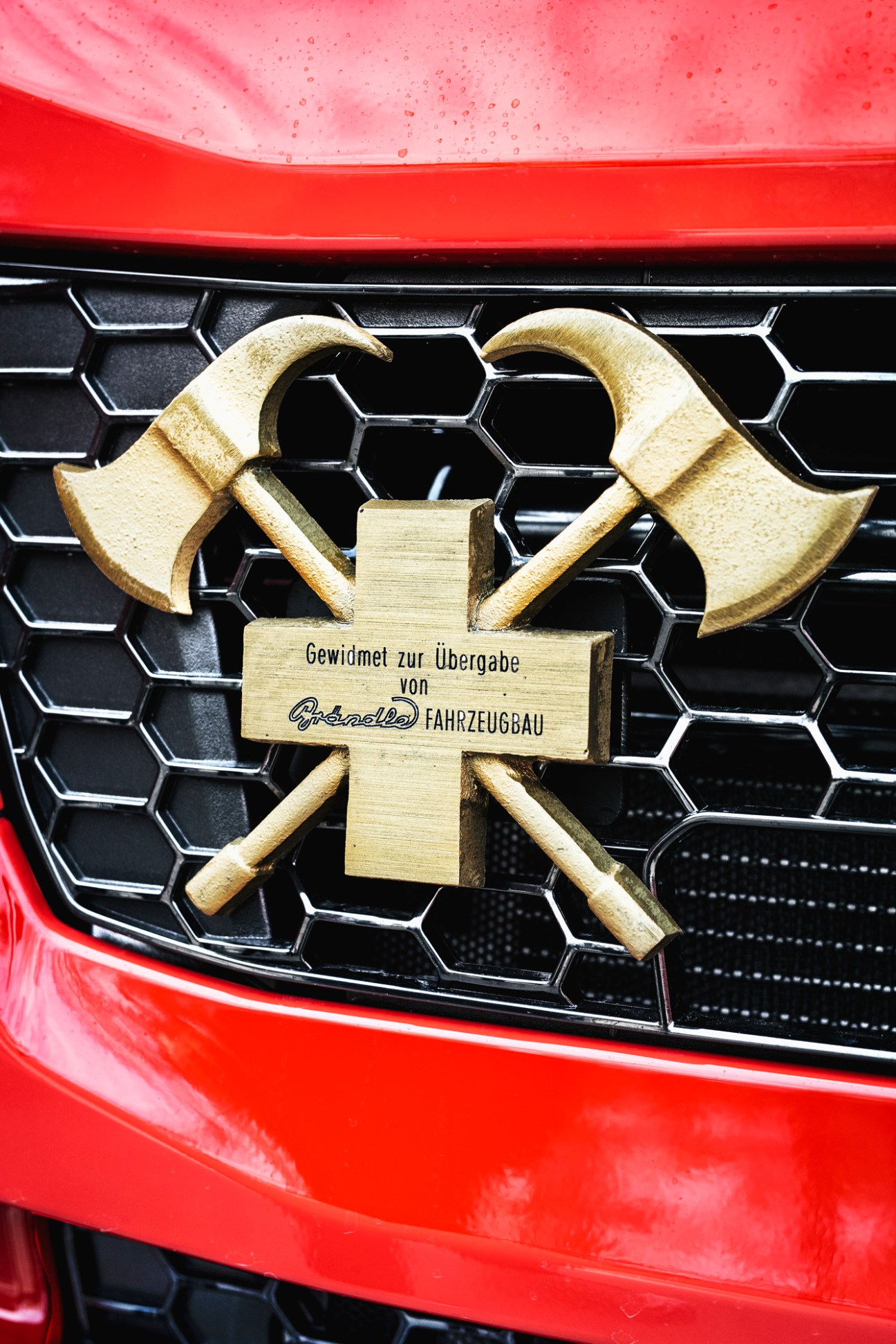 Golden firefighters' cross emblem mounted on black hexagonal grille of red Brändle fire engine
