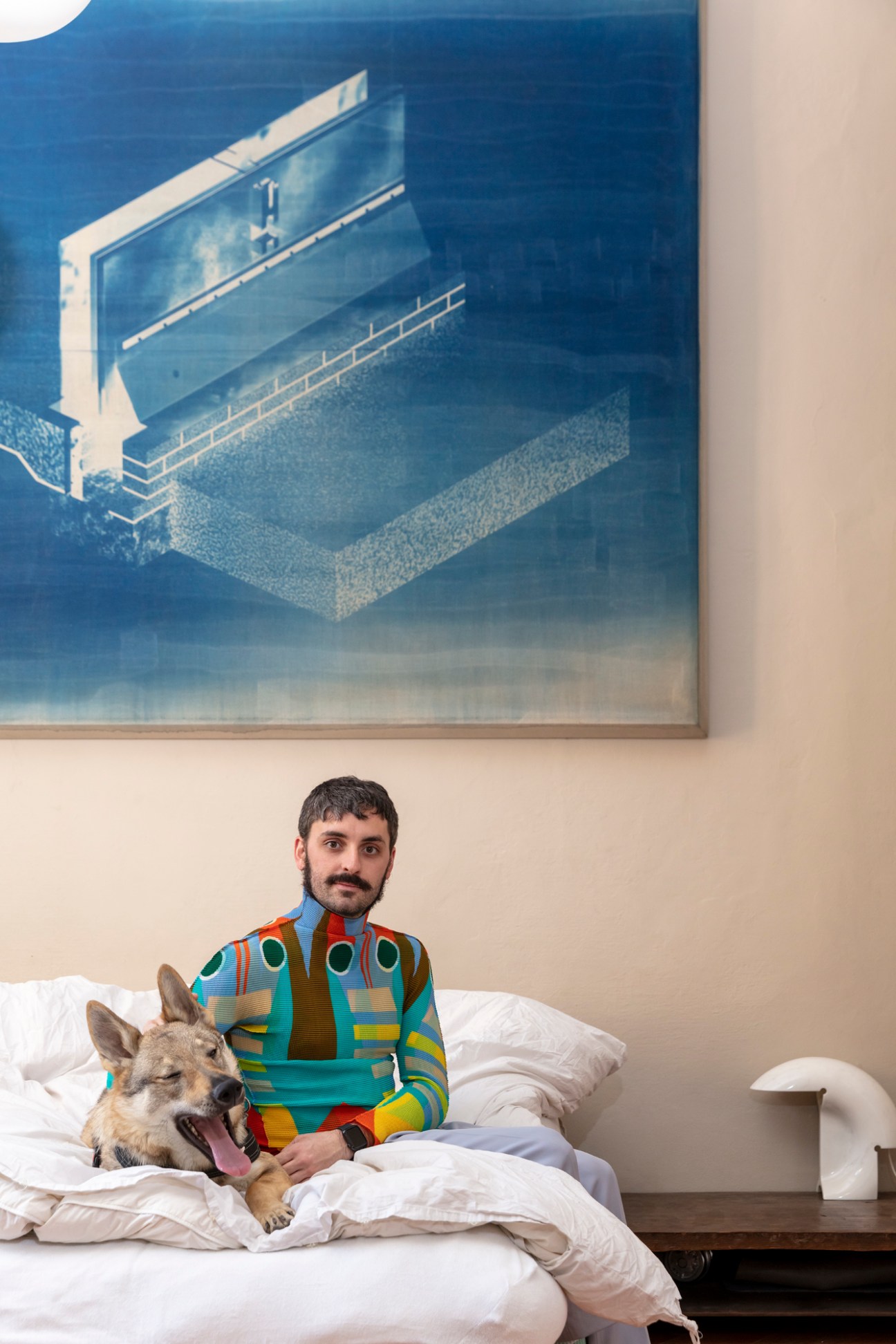 Edoardo Monti sitting on white bed with dog, wearing colorful striped sweater beneath blue artwork