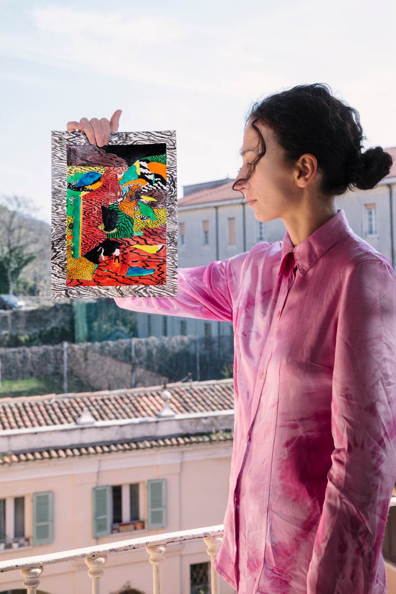 Woman in pink shirt holding up colorful artwork with patterns and birds on balcony overlooking buildings